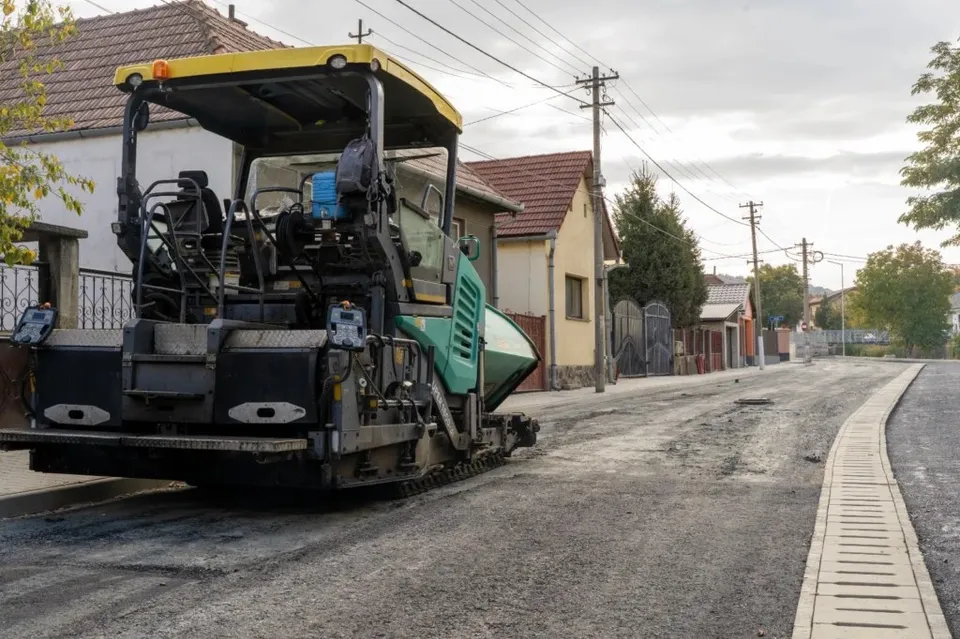 O mașină de asfaltat neagră și verde stă pe un drum de pietriș lângă un trotuar nou și case de proprietate.
