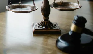 Pe o masă de lemn sunt o balanță metalică și un ciocan de judecată negru, ambele în plan apropiat.