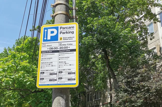 Un panou informativ de parcare galben este montat pe un stâlp de beton lângă copaci verzi și o clădire.