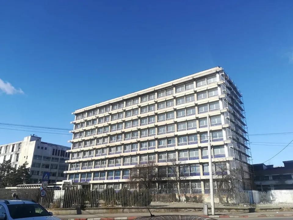 O clădire înaltă cu multe ferestre și fațadă albă se ridică sub un cer albastru senin.