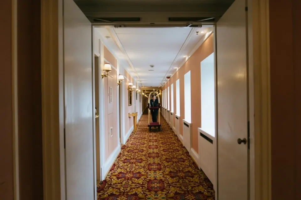 O persoană împinge un cărucior de bagaje pe un coridor lung cu mochetă decorată și uși de culoare roz deschis.