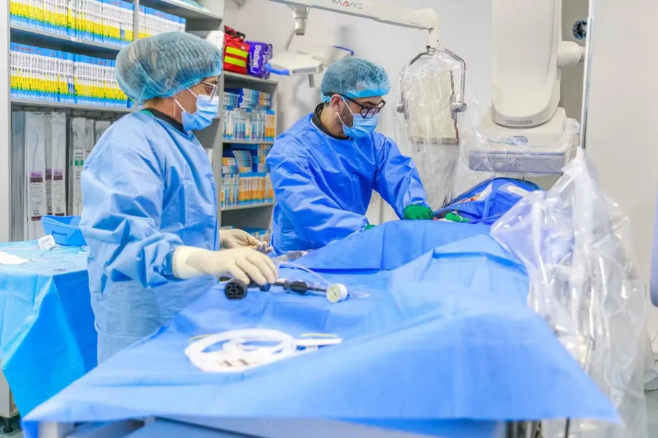 Două persoane purtând halate și măști de protecție albastre lucrează lângă un echipament medical acoperit cu material steril albastru.