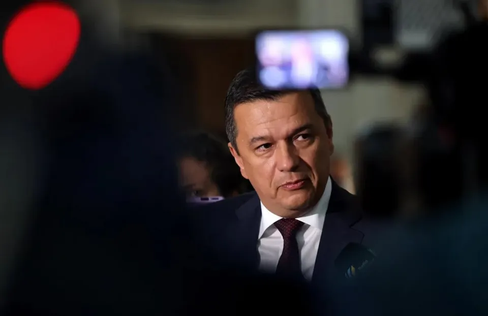 Un bărbat cu părul scurt, purtând sacou închis la culoare, cămașă albă și cravată purpurie, este surprins într-un cadru cu elemente de fundal neclare.