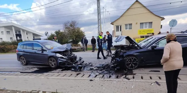 Două autoturisme cu capote deschise sunt oprite pe un drum, în timp ce mai mulți oameni stau în apropierea vehiculelor avariate.