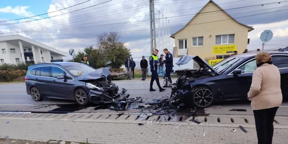 Două autoturisme cu capote deschise sunt oprite pe un drum, în timp ce mai mulți oameni stau în apropierea vehiculelor avariate.