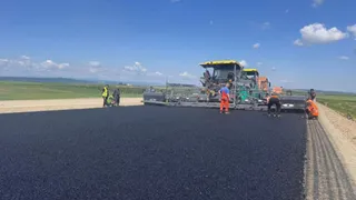 O mașină de asfalt și mai mulți muncitori în haine reflectorizante lucrează pe o porțiune de drum proaspăt turnat sub un cer senin.