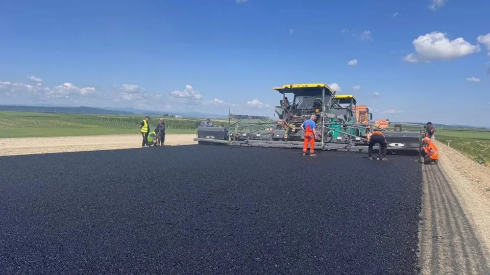 O mașină de asfalt și mai mulți muncitori în haine reflectorizante lucrează pe o porțiune de drum proaspăt turnat sub un cer senin.