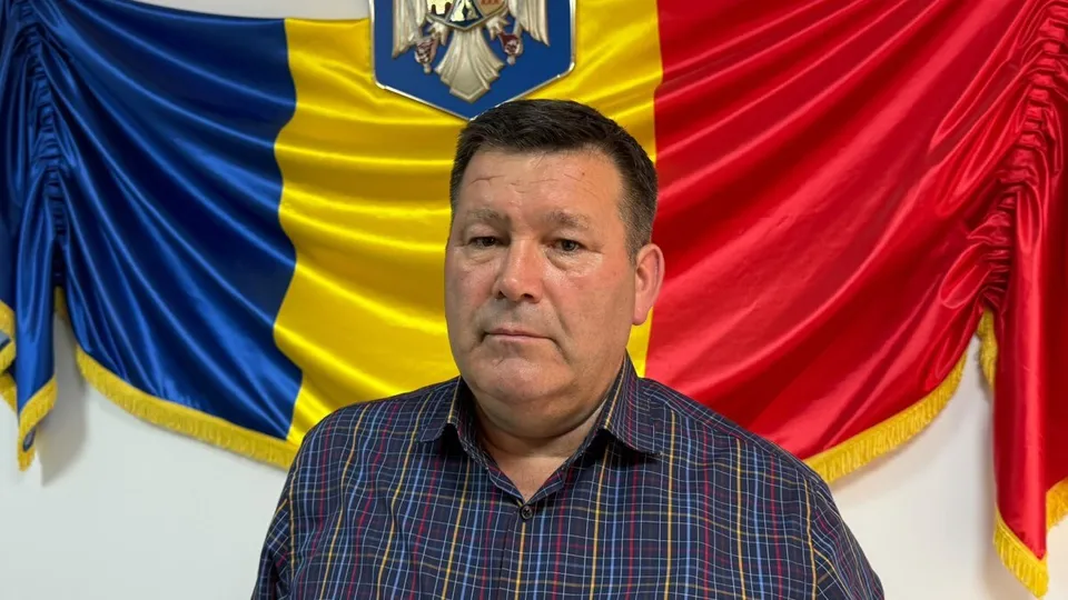 Un bărbat cu cămașă în carouri este poziționat în fața unui steag tricolor pe un fundal alb.