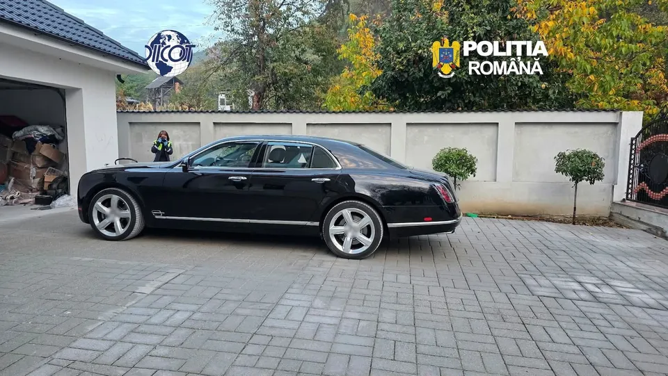 Un sedan negru este parcat pe o curte pavată în fața unui zid alb, lângă o clădire și un arbust mic.