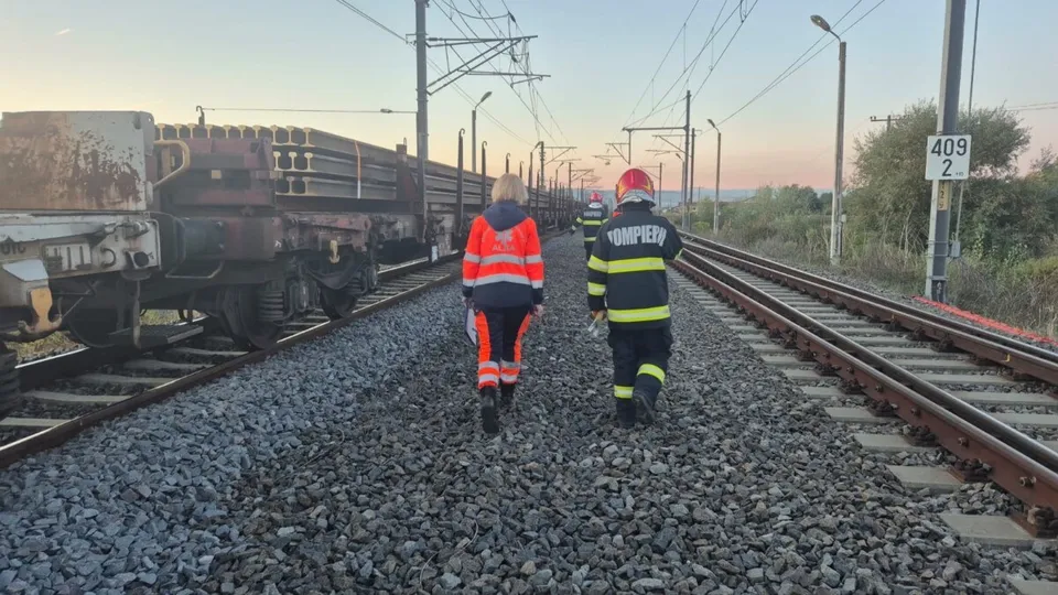 Două persoane purtând veste reflectorizante și echipament de protecție merg pe balastul dintre două căi ferate lângă un tren de marfă încărcat.