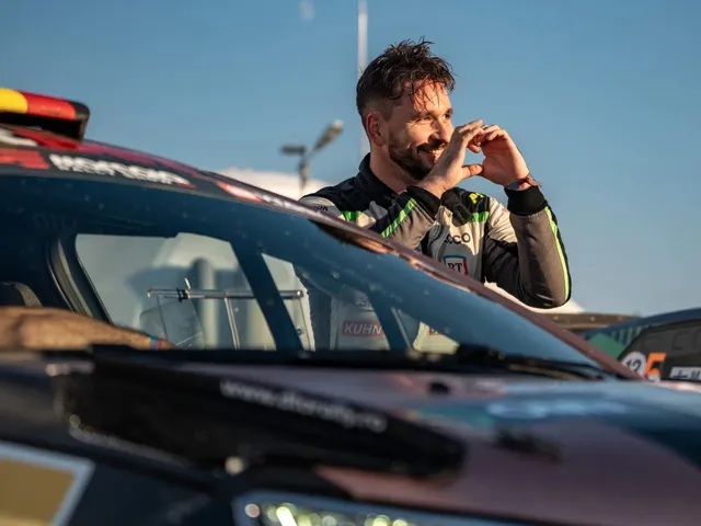 Un bărbat cu barbă și părul scurt, purtând un costum de curse gri și negru, stă lângă un autoturism de competiție sub cerul senin.
