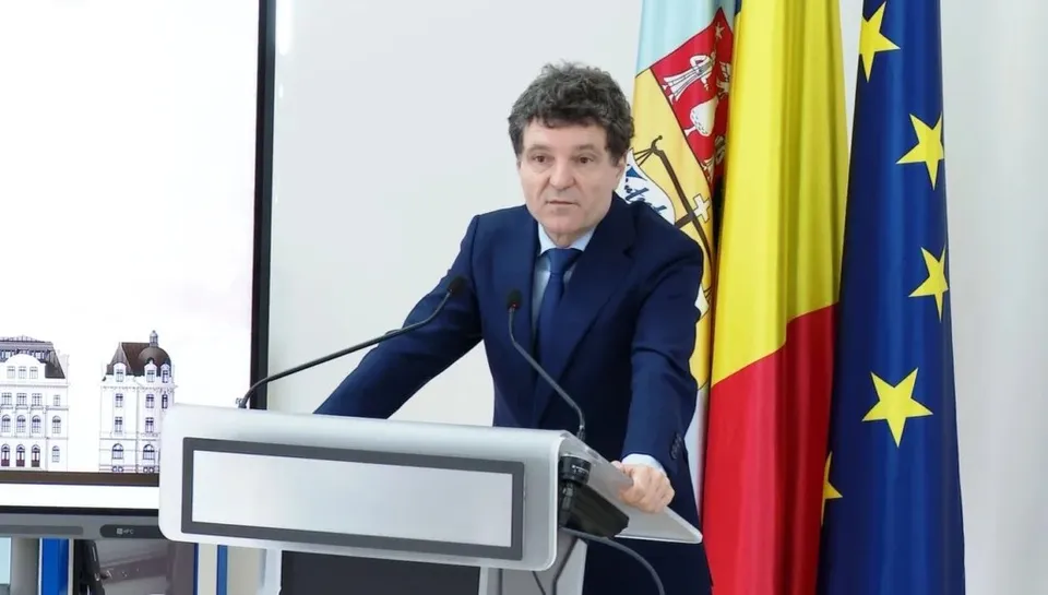 Un bărbat într-un costum albastru stă în spatele unui podium lângă trei steaguri, într-o încăpere cu un ecran mare în fundal.