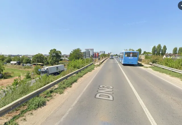 O autostradă cu două benzi de circulație, marcajele DN3 pe asfalt și un autobuz albastru circulând în depărtare sub un cer senin.