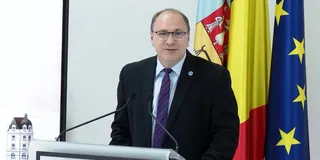 Un bărbat cu ochelari, purtând costum închis la culoare și cravată mov, stă în spatele unui podium lângă trei steaguri.