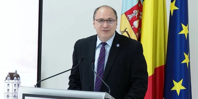 Un bărbat cu ochelari, purtând costum închis la culoare și cravată mov, stă în spatele unui podium lângă trei steaguri.