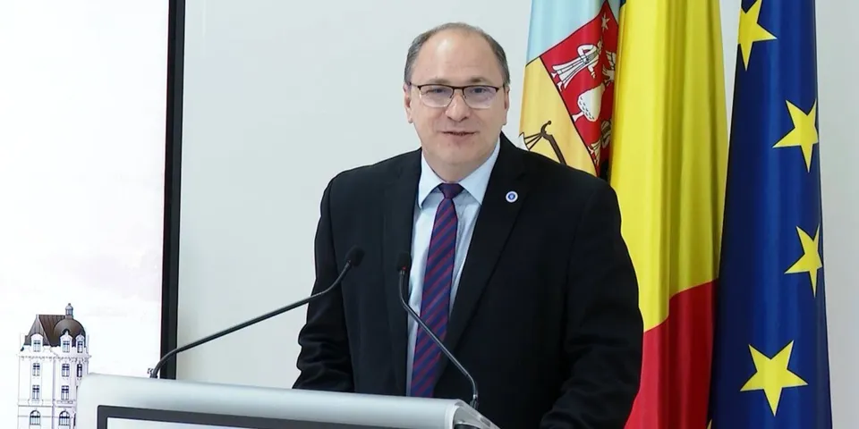 Un bărbat cu ochelari, purtând costum închis la culoare și cravată mov, stă în spatele unui podium lângă trei steaguri.