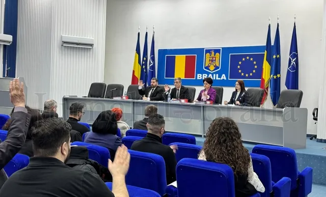 Oamenii stau în scaune albastre într-o sală, în timp ce un grup de persoane este așezat la o masă lungă în fața unor steaguri.