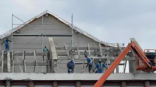 Cinci muncitori cu echipament de protecție lucrează pe un șantier de construcții lângă o structură de beton și o platformă de ridicare portocalie.