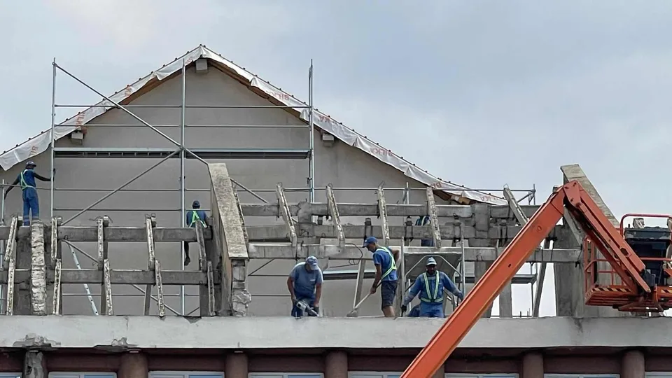 Cinci muncitori cu echipament de protecție lucrează pe un șantier de construcții lângă o structură de beton și o platformă de ridicare portocalie.
