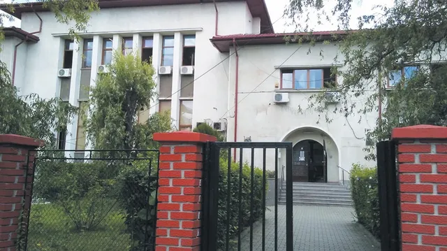 O clădire albă cu ferestre înalte, situată în spatele unui gard de cărămidă roșie și metal negru, înconjurată de vegetație verde.