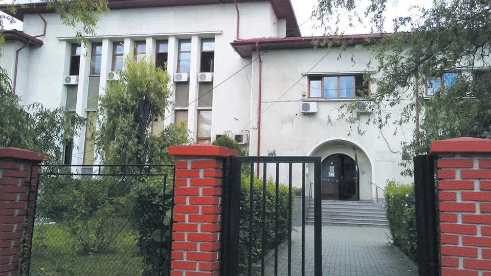 O clădire albă cu ferestre înalte, situată în spatele unui gard de cărămidă roșie și metal negru, înconjurată de vegetație verde.