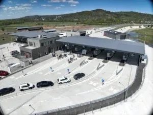 O vedere aeriană a unui punct de frontieră cu mai multe benzi de circulație, mașini parcate și clădiri moderne sub un cer senin.