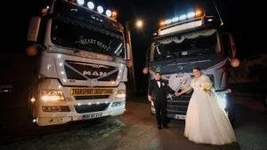 Un bărbat în costum negru și o femeie în rochie de mireasă albă stau în fața a două camioane mari pe un drum noaptea.