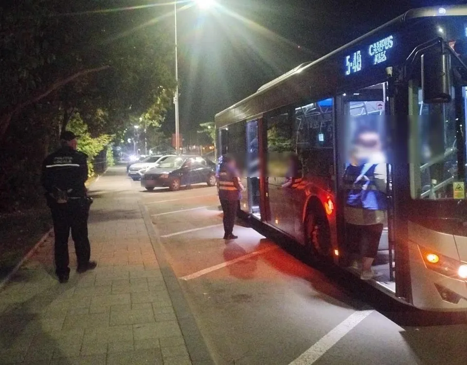 Un agent de poliție stă pe trotuar lângă un autobuz mare parcat pe marginea drumului, în timpul nopții.
