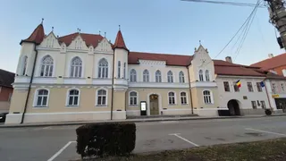 O clădire cu fațadă galbenă și albă prezintă turnuri mici, ferestre arcuate și un steag românesc afișat pe partea dreaptă.