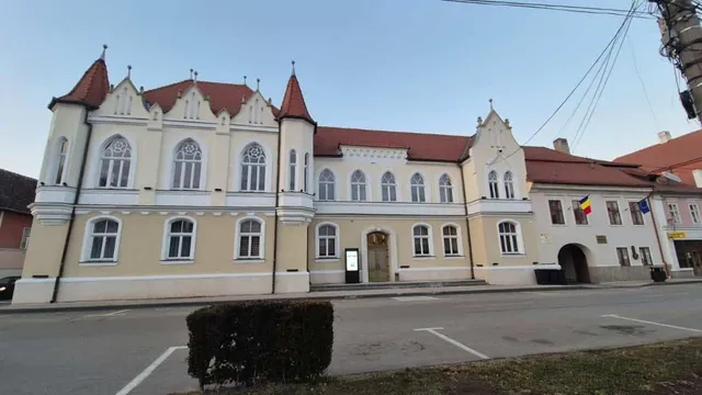O clădire cu fațadă galbenă și albă prezintă turnuri mici, ferestre arcuate și un steag românesc afișat pe partea dreaptă.