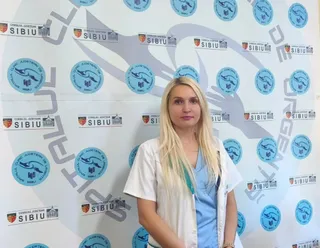 O femeie cu părul blond, purtând halat alb peste un tricou albastru, stă în fața unui fundal alb cu diverse sigilii circulare și dreptunghiulare.