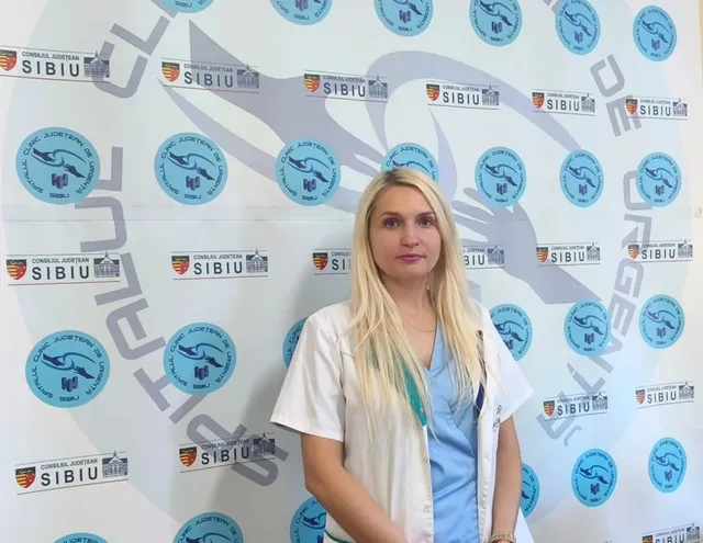 O femeie cu părul blond, purtând halat alb peste un tricou albastru, stă în fața unui fundal alb cu diverse sigilii circulare și dreptunghiulare.