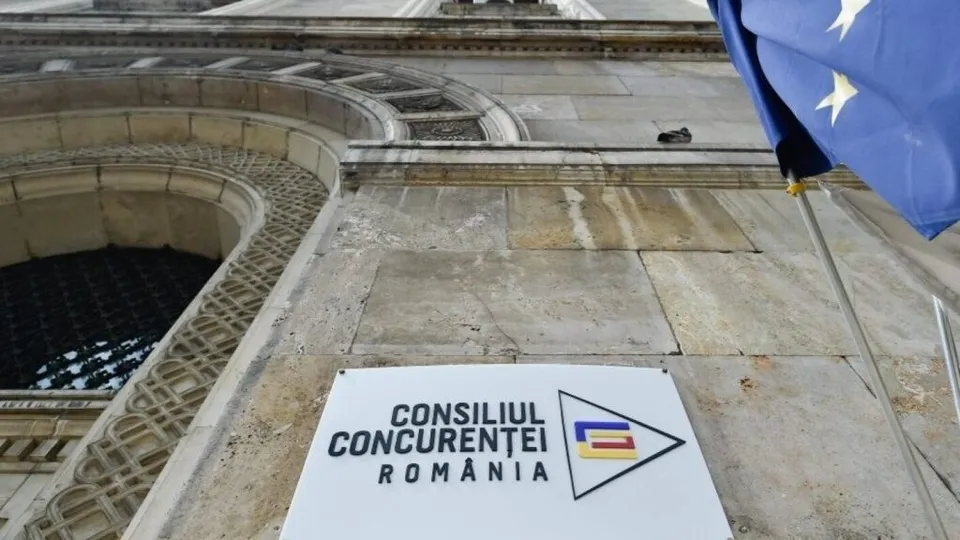 Un panou alb cu textul Consiliul Concurenței România și un logo este montat pe o fațadă de piatră lângă un steag albastru cu stele galbene.