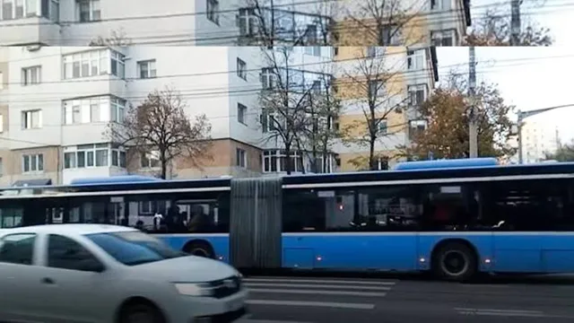 Un autobuz albastru articulat circulă pe un drum lângă un autoturism alb și clădiri de apartamente.