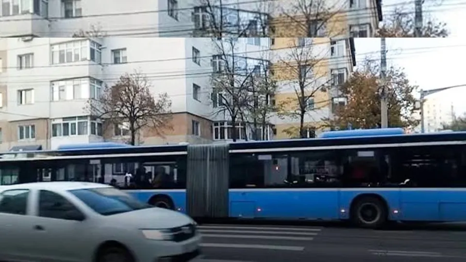 Un autobuz albastru articulat circulă pe un drum lângă un autoturism alb și clădiri de apartamente.