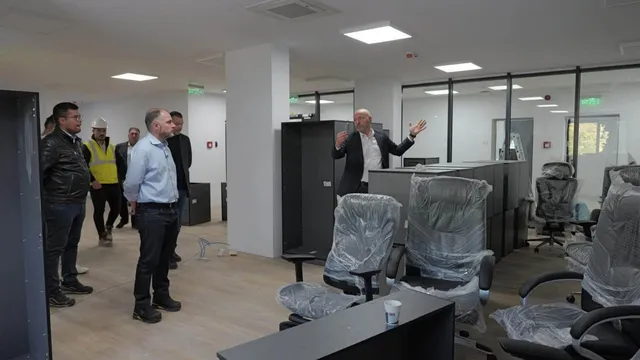 Un grup de bărbați stă într-o încăpere cu birouri, scaune acoperite cu folie și panouri negre de mobilier.