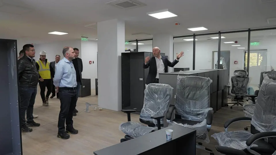 Un grup de bărbați stă într-o încăpere cu birouri, scaune acoperite cu folie și panouri negre de mobilier.