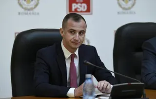Un bărbat cu cămașă albă, cravată roz și sacou închis la culoare stă la o masă lângă un microfon și o sticlă de apă.