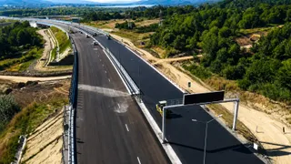 O autostradă proaspăt asfaltată cu linii albe parțial desenate trece printr-un peisaj cu vegetație verde și terenuri de excavare.