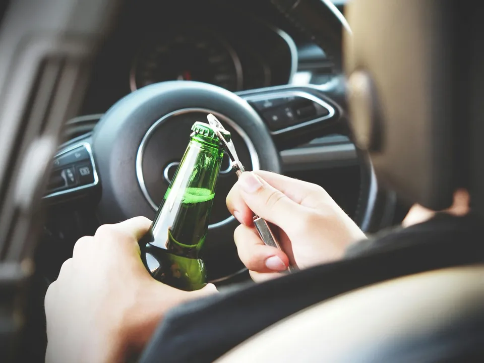 Mâinile unei persoane țin o sticlă de sticlă verde și un decapsator în interiorul unui vehicul, lângă volan.