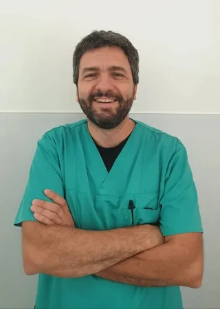 Un bărbat cu barbă și păr scurt, purtând o tunică medicală de culoare verde, stă cu brațele încrucișate în fața unui perete gri și alb.