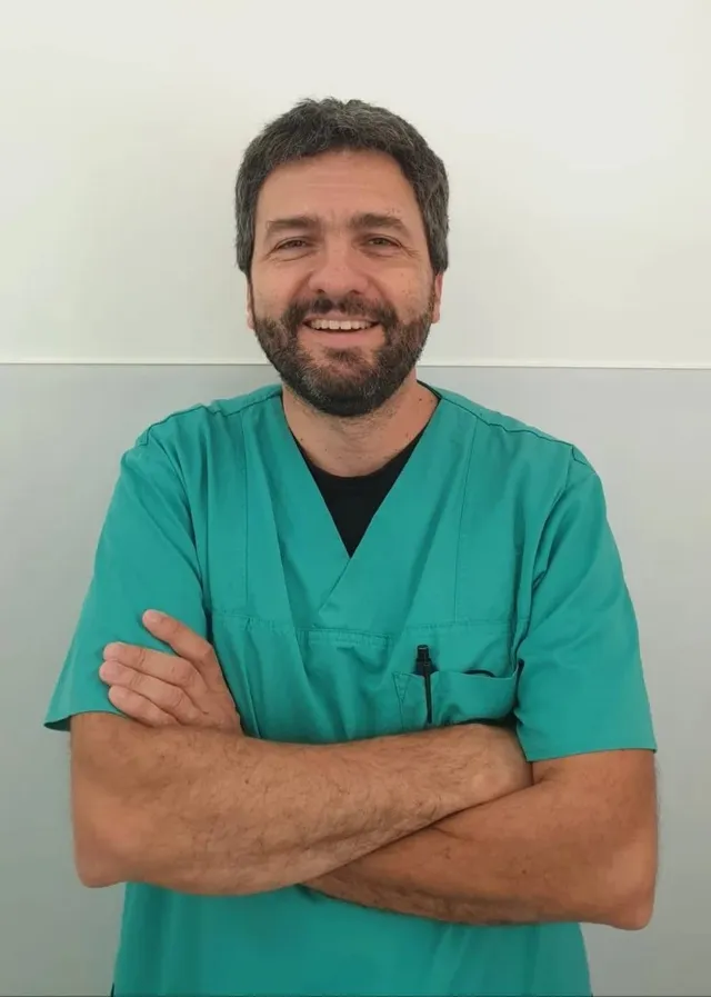Un bărbat cu barbă și păr scurt, purtând o tunică medicală de culoare verde, stă cu brațele încrucișate în fața unui perete gri și alb.