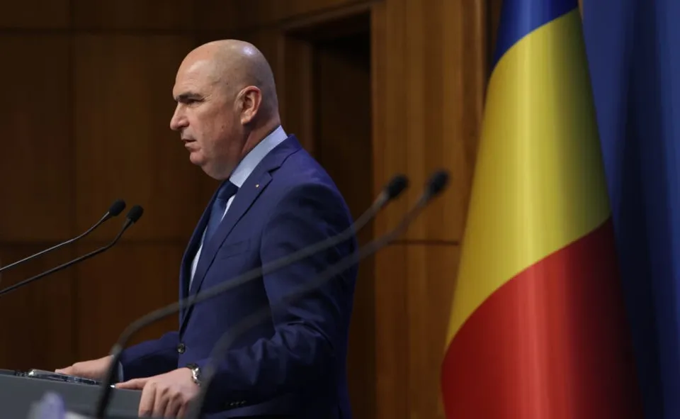 Un bărbat cu capul chel, purtând costum albastru și cravată, stă la un pupitru cu microfoane lângă un steag tricolor.