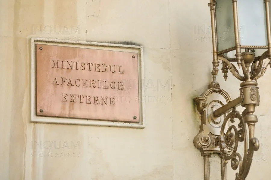 O placă metalică cu textul MINISTERUL AFACERILOR EXTERNE este montată pe un perete deschis lângă un corp de iluminat decorativ din metal.