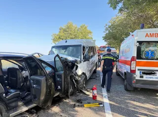 Două vehicule albe și un autoturism negru sunt implicate într-o coliziune pe un drum, lângă o ambulanță și un oficial cu vestă reflectorizantă.