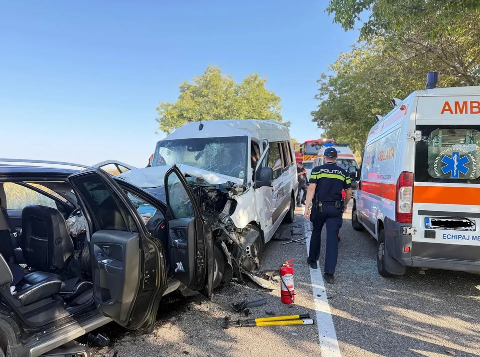 Două vehicule albe și un autoturism negru sunt implicate într-o coliziune pe un drum, lângă o ambulanță și un oficial cu vestă reflectorizantă.
