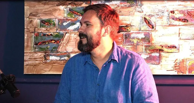Un bărbat cu barbă și cămașă albastră stă în fața unui tablou abstract cu texturi colorate.