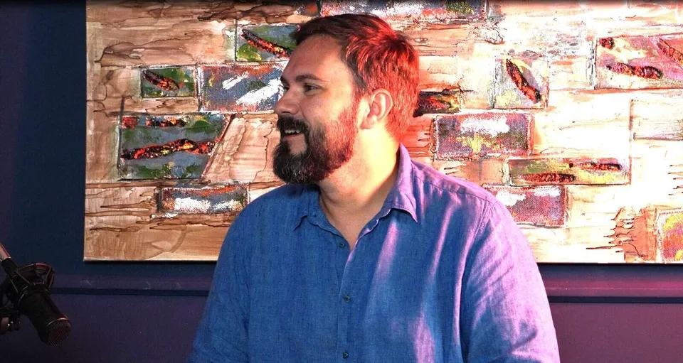 Un bărbat cu barbă și cămașă albastră stă în fața unui tablou abstract cu texturi colorate.