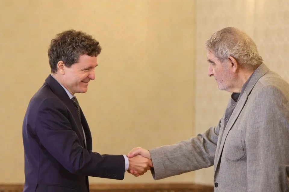 Un bărbat într-un costum închis se strânge în mână cu un bărbat într-un sacou gri în fața unui perete de culoare deschisă.