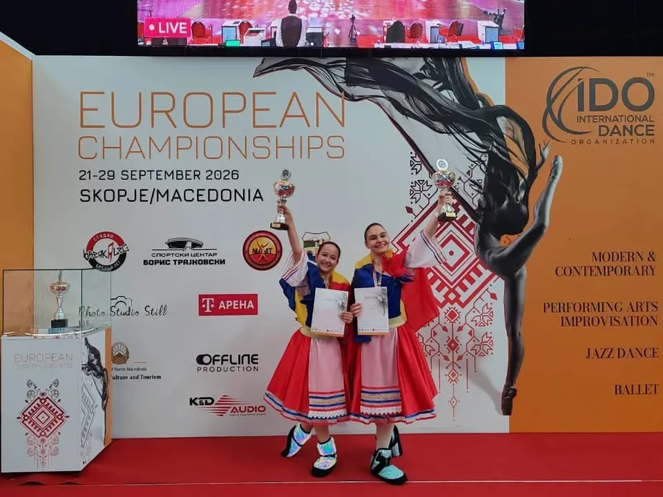 Două fete în costume tradiționale colorate țin trofee și certificate, stând pe un podium în fața unui panou cu inscripții și grafică de dans.